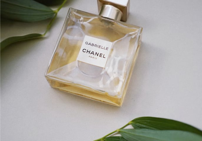 Gabrielle Essence Eau De Parfum Laying flat on a table with green leaves above and below it